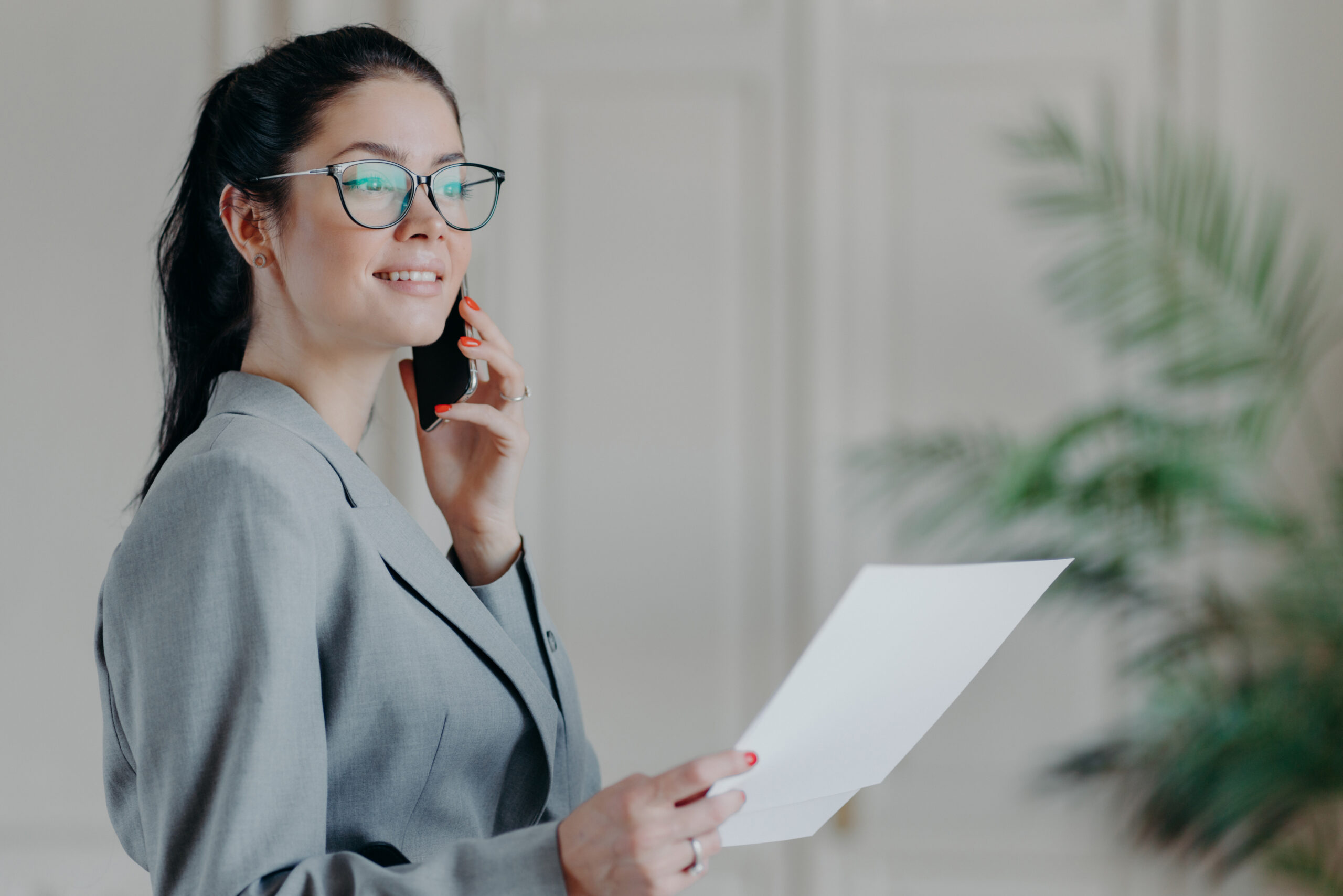 Photo of elegant brunette woman in formal clothes works with papers in office, discusses business issues via smartphone with colleague. Female manager checks information from documents, makes research